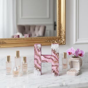 May include: A decorative letter "H" with a pink and gold marbled design, accented with clear crystals. Several perfume bottles and a small orchid plant are arranged on a marble surface. A gold-framed mirror is in the background.