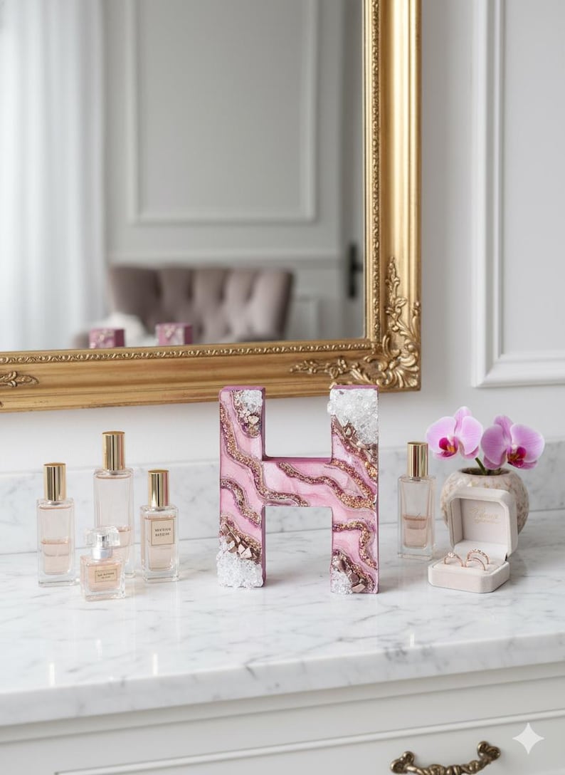 May include: A decorative letter "H" with a pink and gold marbled design, accented with clear crystals. Several perfume bottles and a small orchid plant are arranged on a marble surface. A gold-framed mirror is in the background.
