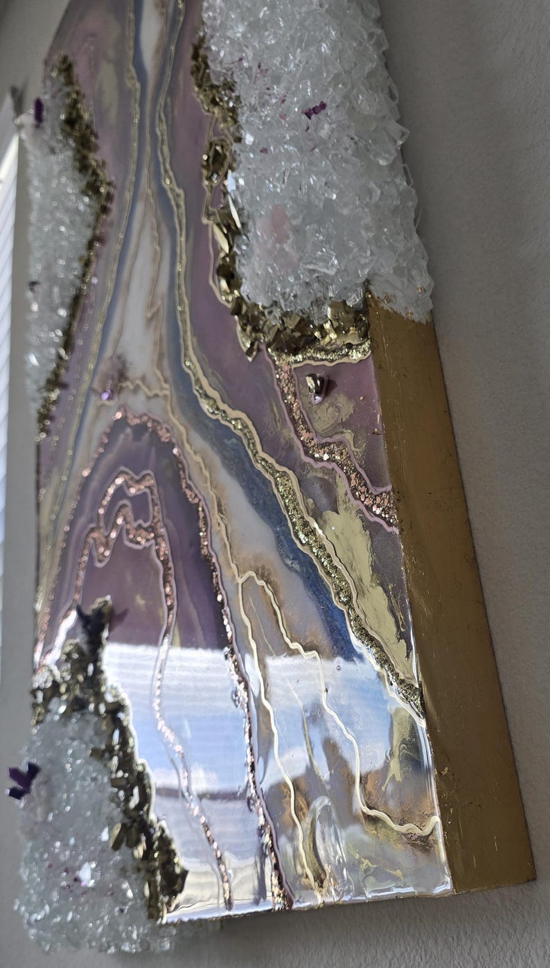 Może przedstawiać: Abstrakcyjne dzieło sztuki z żywicy z pozłacaną krawędzią. Dzieło zawiera warstwy lawendy, bieli i złota, z wbudowanymi kryształami i brokatowymi akcentami. Projekt przypomina geodę lub krajobraz.