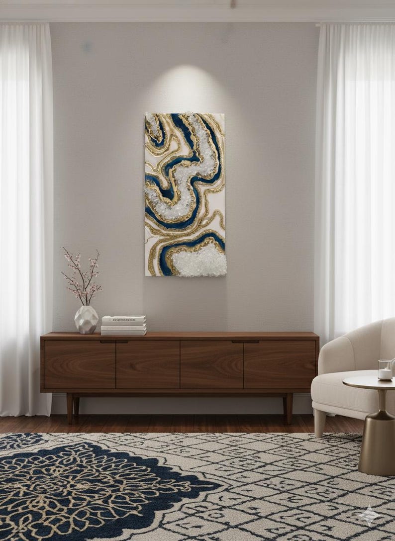 Op de afbeelding: Abstracte wandkunst met een wervelend ontwerp in wit, marineblauw en goud. Het kunstwerk is gemonteerd op een witte achtergrond en hangt boven een houten kast. De kamer bevat ook een patroonkleed, een witte stoel en transparante gordijnen.