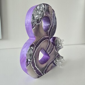 May include: A decorative ampersand sculpture in shades of purple, silver, and gray. The sculpture has a geode-like appearance with crystalline accents and glittery details. The ampersand is a unique home decor item.