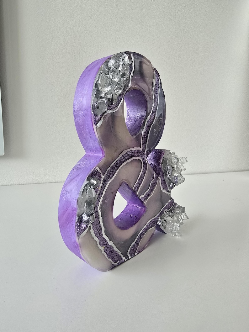 May include: A decorative ampersand sculpture in shades of purple, silver, and gray. The sculpture has a geode-like appearance with crystalline accents and glittery details. The ampersand is a unique home decor item.