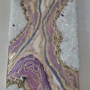 Może przedstawiać: Abstrakcyjna grafika ścienna z motywem geody w odcieniach fioletu, złota i bieli. Grafika zawiera kryształowe akcenty i brokat, tworząc efekt tekstury i wymiaru. Zamontowana na drewnianym panelu.