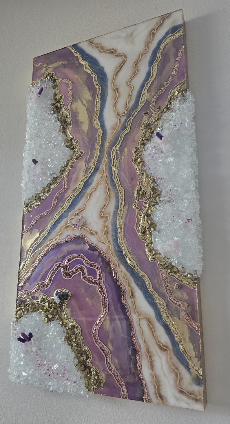 Może przedstawiać: Abstrakcyjna grafika ścienna z motywem geody w odcieniach fioletu, złota i bieli. Grafika zawiera kryształowe akcenty i brokat, tworząc efekt tekstury i wymiaru. Zamontowana na drewnianym panelu.