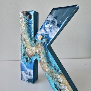 May include: A handcrafted, blue resin letter "K" decorated with a beach theme. The letter features embedded seashells, clear crystals, and a wave design. The letter is approximately 10 inches tall.