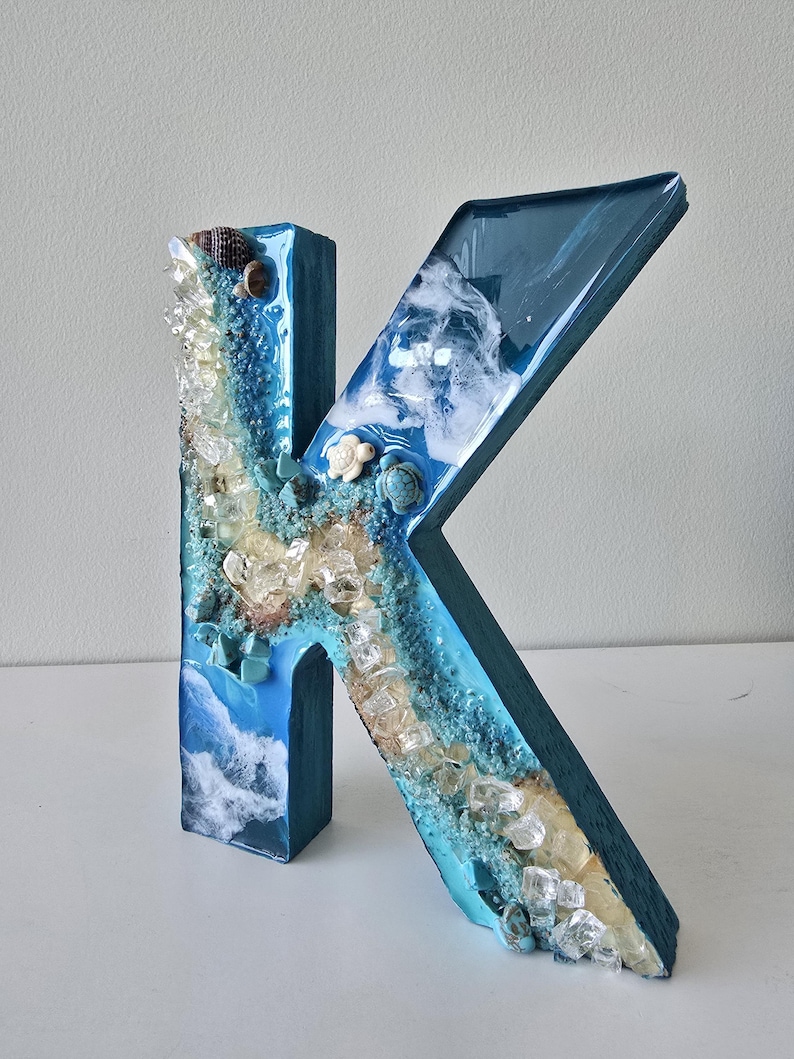 May include: A handcrafted, blue resin letter "K" decorated with a beach theme. The letter features embedded seashells, clear crystals, and a wave design. The letter is approximately 10 inches tall.