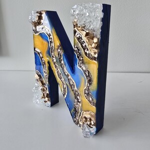 Puede incluir: Una letra "N" decorativa de madera con exterior azul marino. El interior presenta un dise&ntilde;o de resina amarilla y azul, acentuado con purpurina plateada, adornos dorados y detalles cristalinos transparentes. La letra est&aacute; de pie.