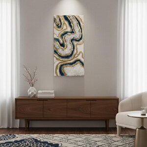 Op de afbeelding: Abstracte wandkunst met een wervelend ontwerp in wit, marineblauw en goud. Het kunstwerk is gemonteerd op een witte achtergrond en hangt boven een houten kast. De kamer omvat ook een patroonkleed, een witte stoel en transparante gordijnen.