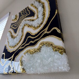 May include: Abstract wall art with a geode design. The artwork features a black, white, and gold colour scheme, with a textured, crystal-like element. The piece is mounted on a square canvas and is displayed on a wall.