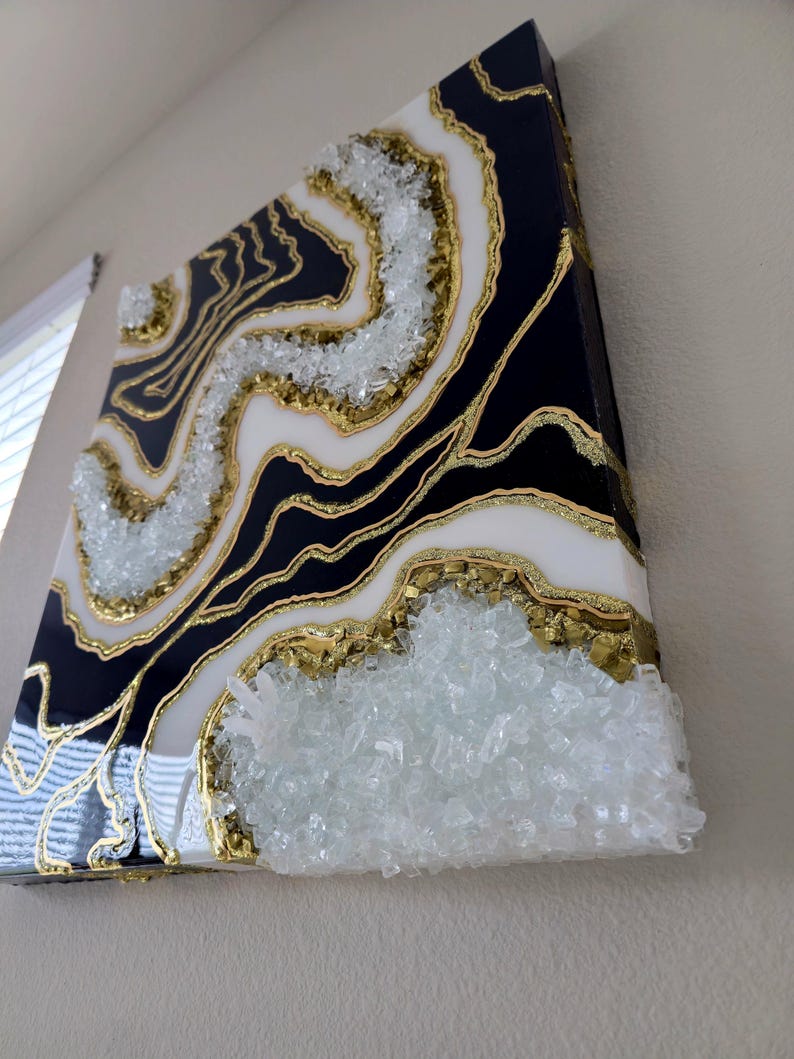 May include: Abstract wall art with a geode design. The artwork features a black, white, and gold colour scheme, with a textured, crystal-like element. The piece is mounted on a square canvas and is displayed on a wall.