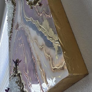 Może przedstawiać: Abstrakcyjna grafika ścienna z warstwowym wzorem w odcieniach lawendy, bieli i złota. Grafika ma żywiczne wykończenie z osadzonymi kryształami i złotymi krawędziami, tworząc efekt tekstury i wymiaru.