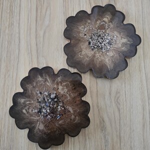 K&ouml;nnte beinhalten: Zwei bl&uuml;tenf&ouml;rmige Untersetzer mit einem dunkelbraunen und beigefarbenen Marmormuster. Die Mitte jedes Untersetzers ist mit silbernem Glitzer verziert. Die Untersetzer sind aus Kunstharz.