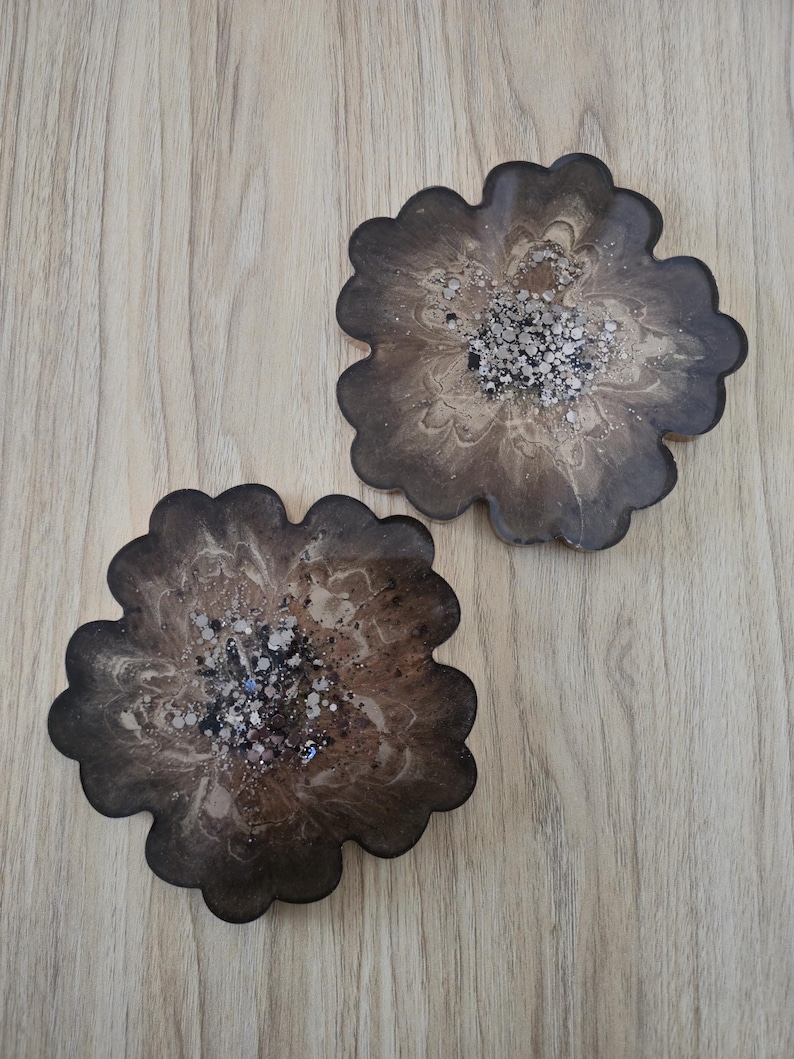 K&ouml;nnte beinhalten: Zwei bl&uuml;tenf&ouml;rmige Untersetzer mit einem dunkelbraunen und beigefarbenen Marmormuster. Die Mitte jedes Untersetzers ist mit silbernem Glitzer verziert. Die Untersetzer sind aus Kunstharz.