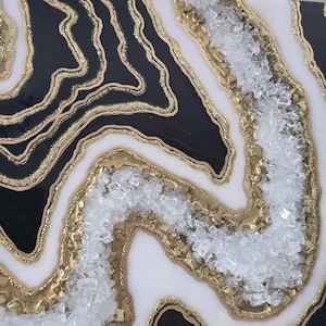 May include: Abstract artwork with a geode design. The piece features black and white sections with gold glitter and gold accents. Clear, crushed glass is embedded in the centre, creating a textured, layered effect. A unique decorative item.