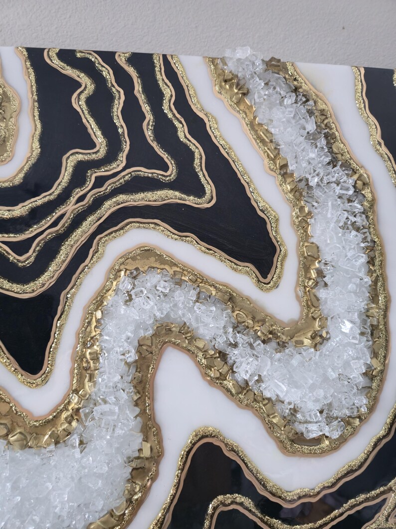 May include: Abstract artwork with a geode design. The piece features black and white sections with gold glitter and gold accents. Clear, crushed glass is embedded in the centre, creating a textured, layered effect. A unique decorative item.