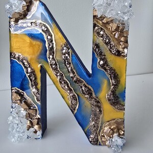 May include: Decorative letter "N" with a blue, yellow, and gold resin design. The letter is embellished with glitter and crystal accents. The letter is a decorative item.
