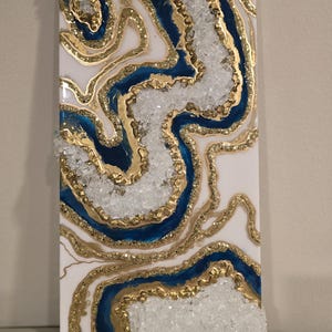 Op de afbeelding: Abstracte wandkunst met een geode-ontwerp. Het kunstwerk heeft een witte achtergrond met blauwe en gouden accenten. Heldere, kristalachtige elementen zijn in het ontwerp verwerkt, wat een textuureffect cre&euml;ert. Het stuk is rechthoekig en op een wit oppervlak gemonteerd.