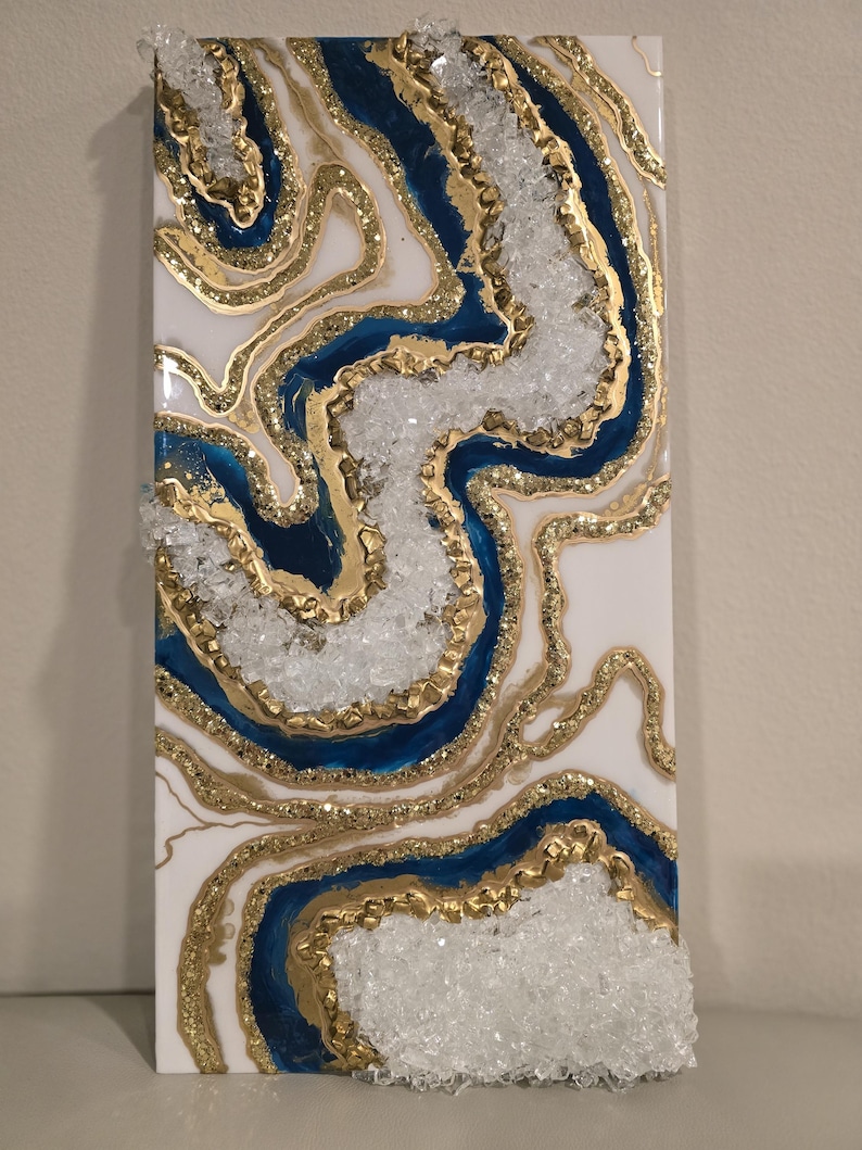 Op de afbeelding: Abstracte wandkunst met een geode-ontwerp. Het kunstwerk heeft een witte achtergrond met blauwe en gouden accenten. Heldere, kristalachtige elementen zijn in het ontwerp verwerkt, wat een textuureffect cre&euml;ert. Het stuk is rechthoekig en op een wit oppervlak gemonteerd.