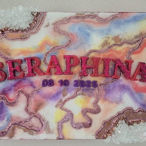 Puede incluir: Una obra de arte cuadrada decorativa con un dise&ntilde;o abstracto colorido, con el nombre "SERAPHINA" en letras de purpurina rosa y la fecha "08 10 2025". La obra est&aacute; adornada con purpurina y detalles de cristal.