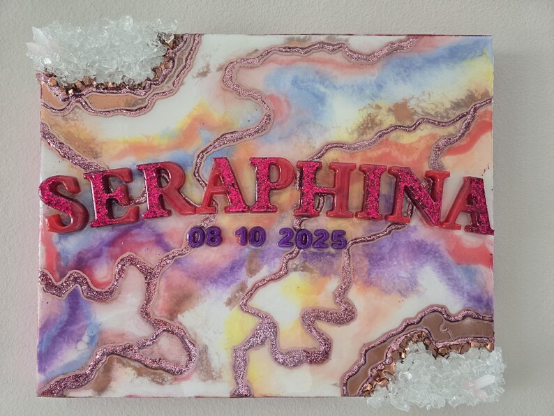 Puede incluir: Una obra de arte cuadrada decorativa con un dise&ntilde;o abstracto colorido, con el nombre "SERAPHINA" en letras de purpurina rosa y la fecha "08 10 2025". La obra est&aacute; adornada con purpurina y detalles de cristal.
