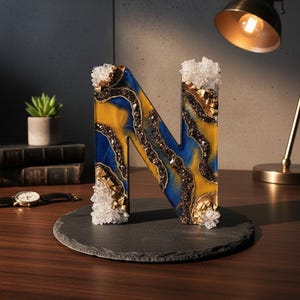 Puede incluir: Una letra "N" decorativa hecha de resina, con un dise&ntilde;o azul y amarillo con detalles marrones y dorados. La letra est&aacute; adornada con adornos transparentes en forma de cristal y se encuentra sobre una base de pizarra gris oscura. La letra mide unos 15 cm de alto.