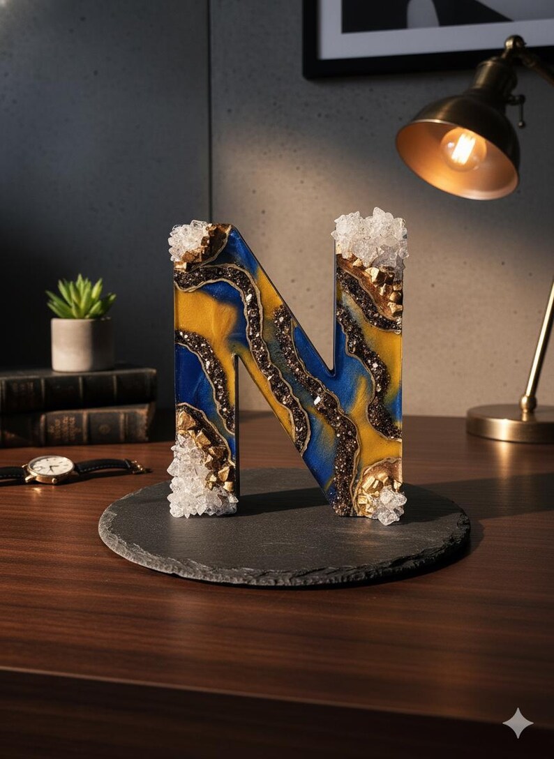 Puede incluir: Una letra "N" decorativa hecha de resina, con un dise&ntilde;o azul y amarillo con detalles marrones y dorados. La letra est&aacute; adornada con adornos transparentes en forma de cristal y se encuentra sobre una base de pizarra gris oscura. La letra mide unos 15 cm de alto.