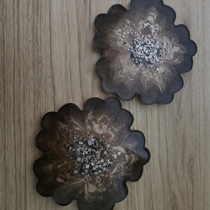 K&ouml;nnte beinhalten: Zwei bl&uuml;tenf&ouml;rmige Untersetzer mit dunkelbraunem, marmoriertem Design. Die Untersetzer haben einen gewellten Rand und ein zentrales Muster aus wei&szlig;en und silbernen Sprenkeln. Die Untersetzer sind aus Kunstharz.
