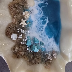 May include: A decorative seashell-shaped dish with a beach scene. The dish features a sandy area with seashells, a starfish, and turquoise stones. Blue and white resin simulates ocean waves.