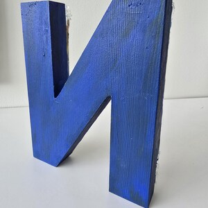 Puede incluir: Una gran letra "N" de madera pintada en un vibrante tono azul. La letra hecha a mano tiene una apariencia r&uacute;stica y texturizada, con vetas de madera visibles y ligeras imperfecciones. La letra es un art&iacute;culo decorativo, adecuado para la decoraci&oacute;n del hogar o regalos personalizados.