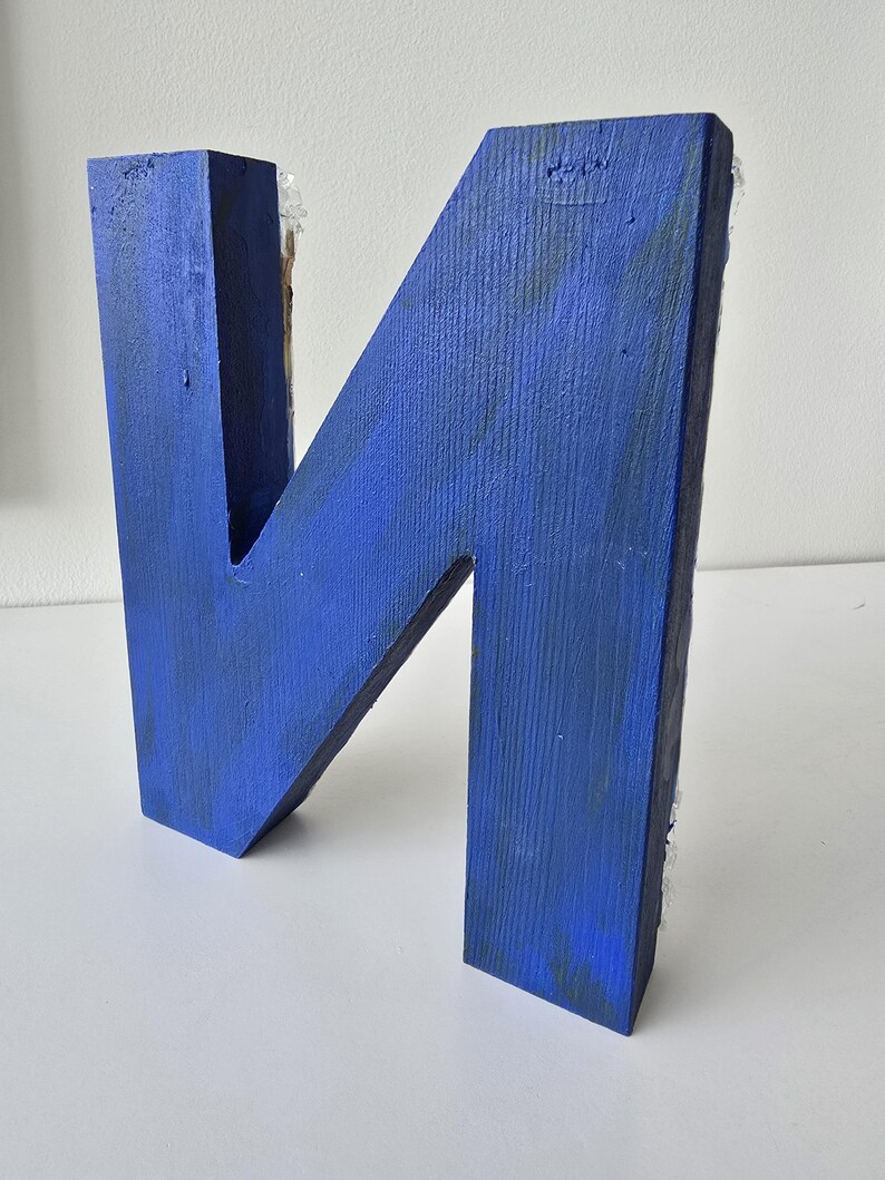 Puede incluir: Una gran letra "N" de madera pintada en un vibrante tono azul. La letra hecha a mano tiene una apariencia r&uacute;stica y texturizada, con vetas de madera visibles y ligeras imperfecciones. La letra es un art&iacute;culo decorativo, adecuado para la decoraci&oacute;n del hogar o regalos personalizados.