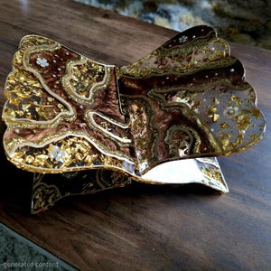 May include: An open, decorative book with a gold and brown design. The book has a layered, textured appearance with gold leaf accents and small white floral embellishments. The edges of the pages have a scalloped design. The book is resting on a dark wooden surface.