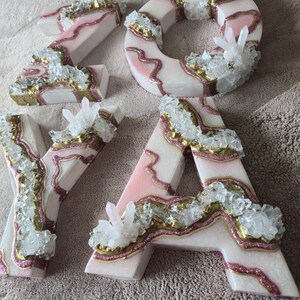May include: Four decorative letters spelling "ZOYA". Each letter is white with pink and gold accents, and embellished with clear crystal clusters. The letters are arranged on a neutral-colored surface.