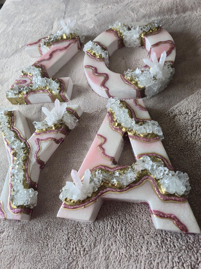 May include: Four decorative letters spelling "ZOYA". Each letter is white with pink and gold accents, and embellished with clear crystal clusters. The letters are arranged on a neutral-colored surface.