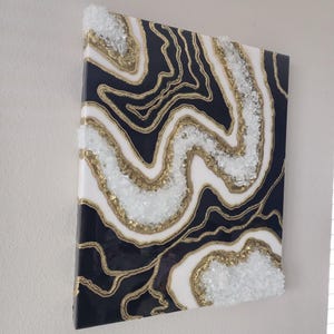May include: Abstract wall art with a geode design. The artwork has a dark navy blue background with white and gold accents. Clear, crushed glass is embedded along the gold lines, creating a textured effect. The piece is mounted on a white wall.