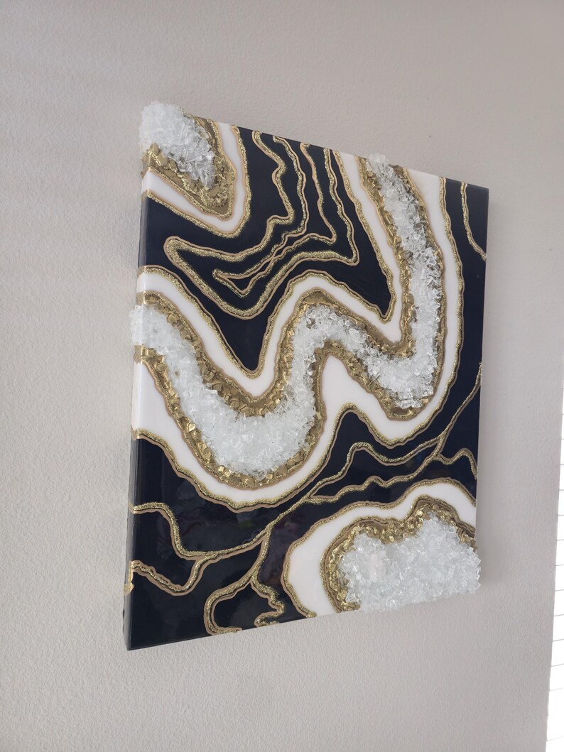 May include: Abstract wall art with a geode design. The artwork has a dark navy blue background with white and gold accents. Clear, crushed glass is embedded along the gold lines, creating a textured effect. The piece is mounted on a white wall.