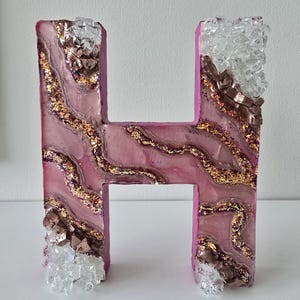 May include: A decorative letter "H" in pink resin with gold glitter and crystal accents. The letter has a geode-like design with layers of pink, gold, and brown. Clear crystals and brown metallic pieces are embedded on the edges.