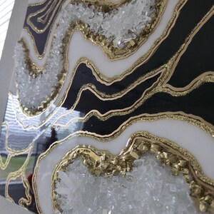 May include: Abstract geode-style wall art in black, white, and gold resin. The artwork includes clear crystal embellishments and gold glitter, creating a textured effect. The piece is mounted on a square canvas, offering a modern decorative element.