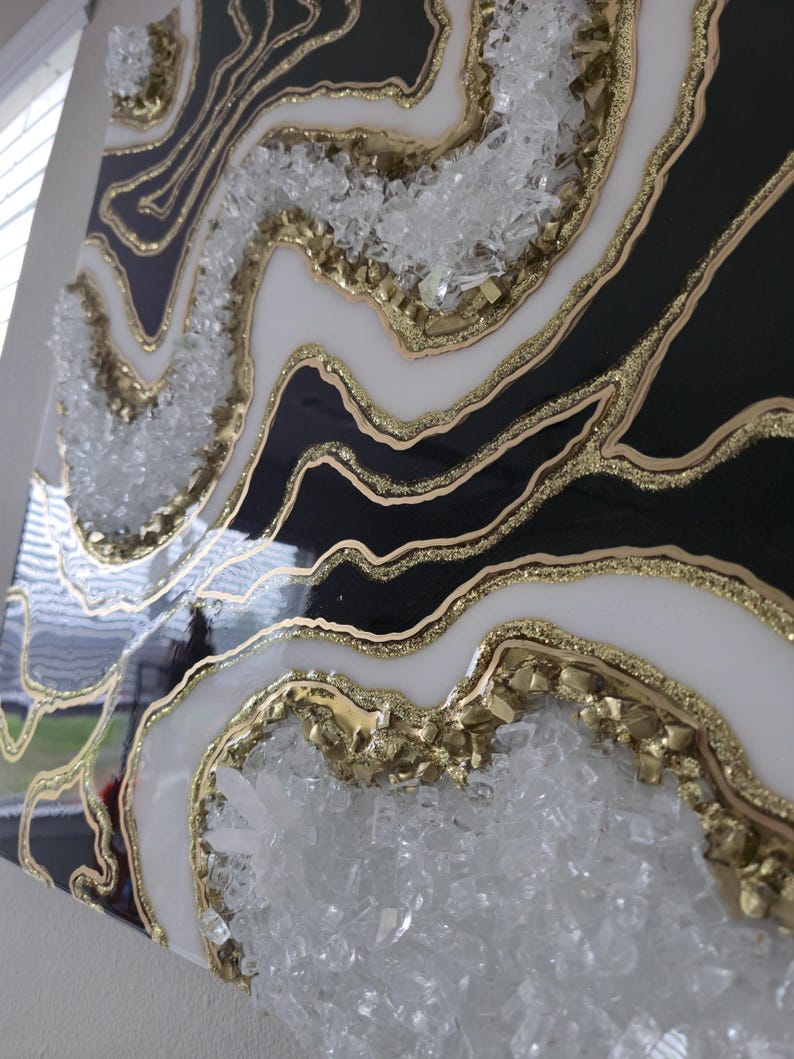 May include: Abstract geode-style wall art in black, white, and gold resin. The artwork includes clear crystal embellishments and gold glitter, creating a textured effect. The piece is mounted on a square canvas, offering a modern decorative element.