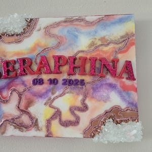 Puede incluir: Una obra de arte cuadrada con un dise&ntilde;o abstracto colorido en tonos rosa, morado y amarillo. El nombre "SERAPHINA" se muestra en letras rosas brillantes en relieve, junto con la fecha "08 10 2025". Acentos de cristal transparente adornan los bordes.