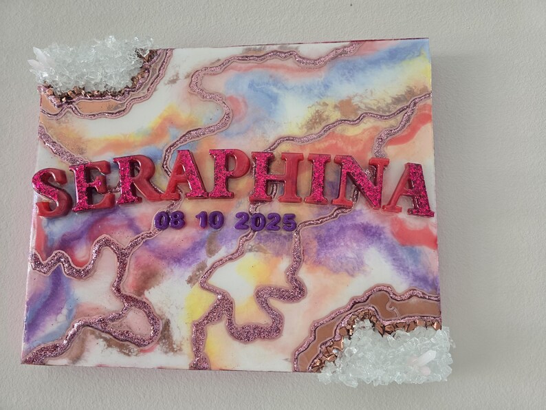 Puede incluir: Una obra de arte cuadrada con un dise&ntilde;o abstracto colorido en tonos rosa, morado y amarillo. El nombre "SERAPHINA" se muestra en letras rosas brillantes en relieve, junto con la fecha "08 10 2025". Acentos de cristal transparente adornan los bordes.