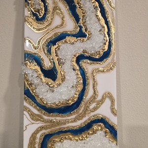 Op de afbeelding: Abstracte wandkunst met een witte achtergrond, blauwe en gouden accenten en heldere kristalachtige elementen. Het ontwerp bevat vloeiende lijnen en texturen, wat een uniek visueel effect cre&euml;ert. Het kunstwerk is rechthoekig en waarschijnlijk op basis van hars.