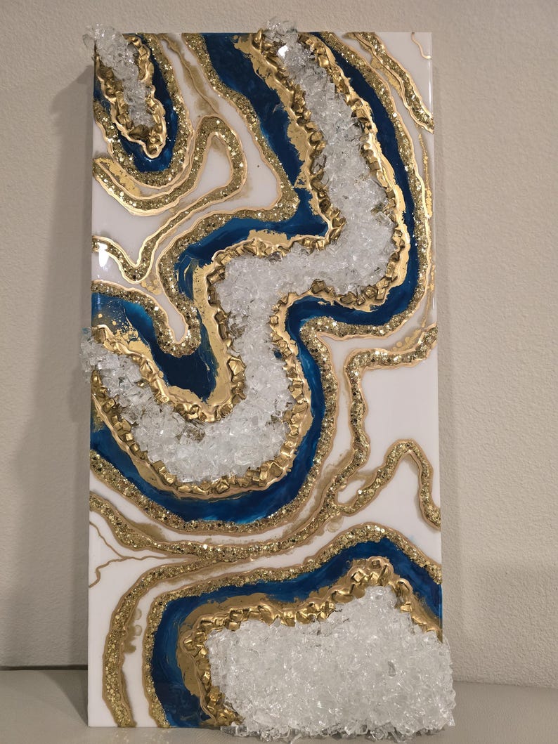 Op de afbeelding: Abstracte wandkunst met een witte achtergrond, blauwe en gouden accenten en heldere kristalachtige elementen. Het ontwerp bevat vloeiende lijnen en texturen, wat een uniek visueel effect cre&euml;ert. Het kunstwerk is rechthoekig en waarschijnlijk op basis van hars.