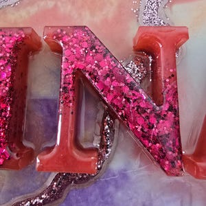 Puede incluir: Primer plano de letras de resina que deletrean una palabra, con la letra "N" en foco. Las letras son de un rojo vibrante, llenas de purpurina rosa. El fondo presenta una mezcla de colores, incluyendo rosa, morado y acentos de purpurina plateada.