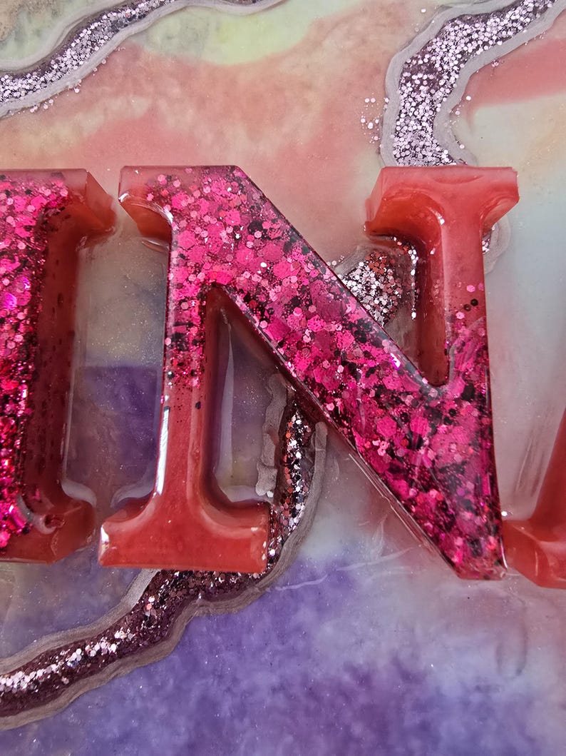 Puede incluir: Primer plano de letras de resina que deletrean una palabra, con la letra "N" en foco. Las letras son de un rojo vibrante, llenas de purpurina rosa. El fondo presenta una mezcla de colores, incluyendo rosa, morado y acentos de purpurina plateada.