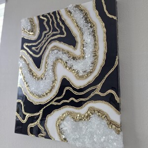 May include: Abstract wall art with a geode design in black, white, and gold. The artwork features layers of black and white with gold accents and clear crystal-like embellishments, creating a textured effect.