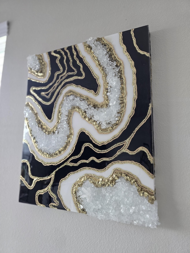 May include: Abstract wall art with a geode design in black, white, and gold. The artwork features layers of black and white with gold accents and clear crystal-like embellishments, creating a textured effect.
