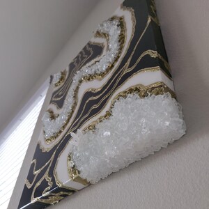 May include: Abstract wall art with a geode design. The artwork features a black and white marbled pattern with gold accents and clear crystal embellishments. The piece is mounted on a square canvas.