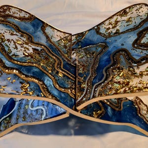 May include: A decorative wooden book stand with a unique design. The stand features a blue and white marbled pattern with gold accents. The edges are outlined in gold, and the overall shape resembles a stylized open book.