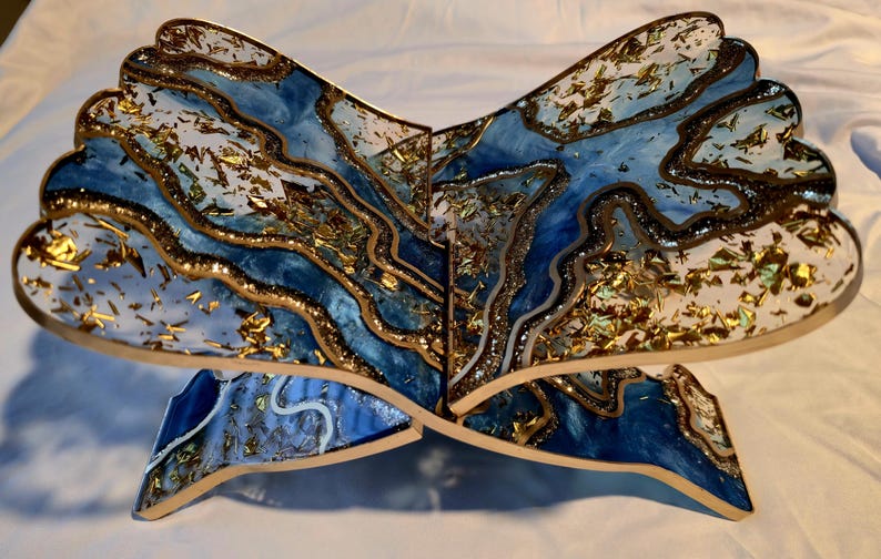 May include: A decorative wooden book stand with a unique design. The stand features a blue and white marbled pattern with gold accents. The edges are outlined in gold, and the overall shape resembles a stylized open book.