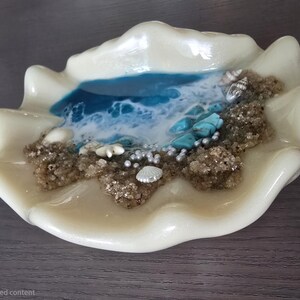 May include: An off-white clam shell-shaped dish featuring a miniature beach scene. It has a blue and white resin ocean, sand-textured areas, seashells, turquoise stones, and pearl beads. The dish is set on a dark surface, creating a coastal theme.